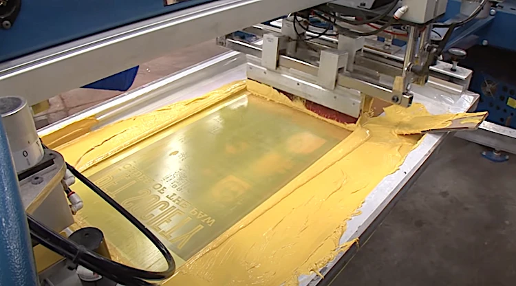 Automated screen printing machine in factory squeegeeing yellow paint through mesh onto substrate.